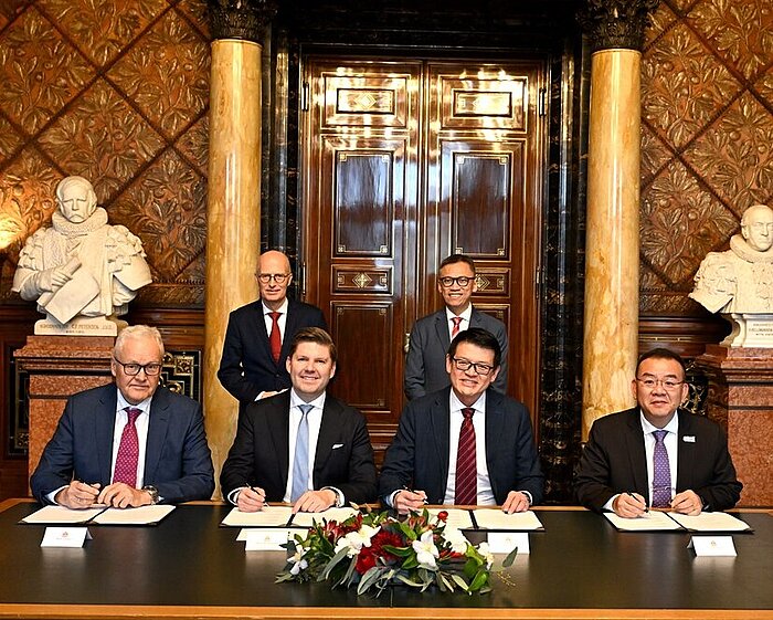 Fünf Personen sitzen an einem Tisch und unterzeichnen Dokumente, dahinter zwei stehende Personen, prunkvoller Raum mit Marmorsäulen und Büsten.