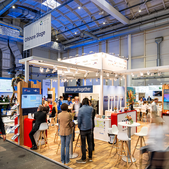 Messestand auf der WindEnergy Hamburg mit Besucherinnen und Besuchern im Gesprächs- und Networkingbereich. Über dem Stand hängt ein Schild mit der Aufschrift „Offshore Stage“. Der offen gestaltete Ausstellungsbereich mit Sitzgelegenheiten, Informationsdisplays und Exponaten zeigt die lebhafte Atmosphäre der Fachmesse.
