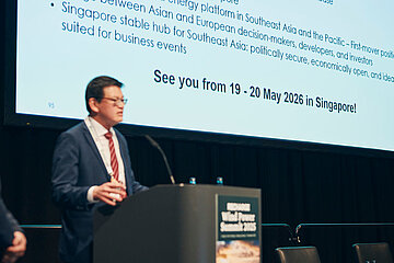 Eine Person im Anzug spricht auf einer Konferenzbühne vor einer großen Präsentationsfolie, die ein Ereignis vom 19.–20. Mai 2026 in Singapur ankündigt.