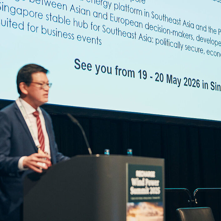 Eine Person im Anzug spricht auf einer Konferenzbühne vor einer großen Präsentationsfolie, die ein Ereignis vom 19.–20. Mai 2026 in Singapur ankündigt.