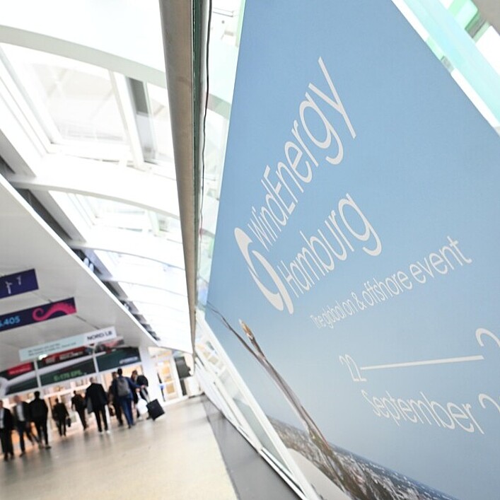 Große Werbefläche mit dem Schriftzug „WindEnergy Hamburg, the global on & offshore event, 22–25 September 2026“ in einem hellen Messegang, während im Hintergrund Personen entlang des Ganges gehen.