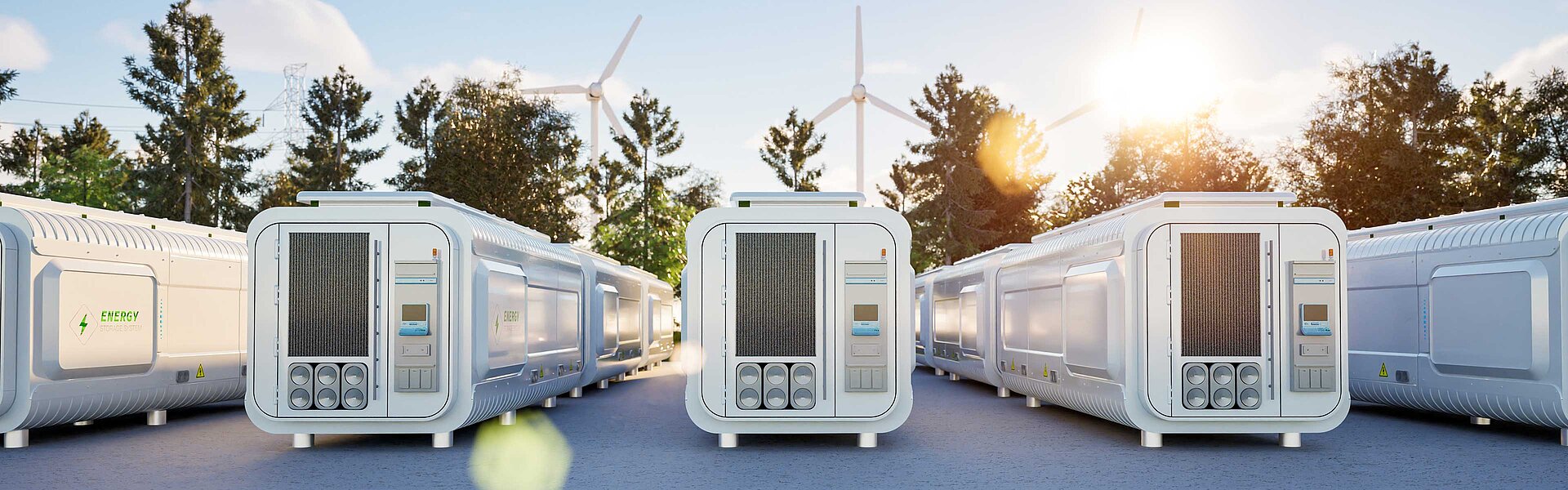 Mehrere Energiespeicher stehen aufgereiht nebeneinander. Hinter den Containern sind blauer Himmel und Windräder zu sehen.