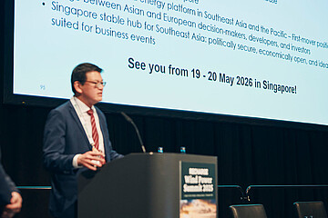 Eine Person im Anzug spricht auf einer Konferenzbühne vor einer großen Präsentationsfolie, die ein Ereignis vom 19.–20. Mai 2026 in Singapur ankündigt.