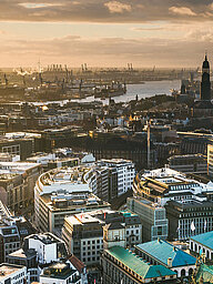 Keyvisual des RECHARGE WindPower Summit. Blick auf Hamburg mit der Hauptkirche St. Michaelis, der Elbphilharmonie, dem Hamburger Hafen und dem Rathaus. Im Hintergrund sind Windräder zu sehen.